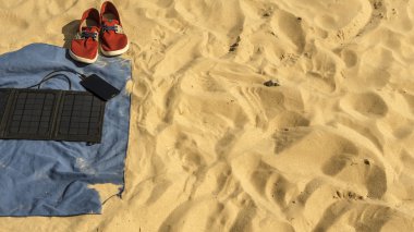 in the sand on the beach are: towel, sneakers, a phone that is charged from a solar panel battery.