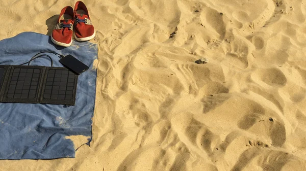 В піску на пляжі є: рушник, кросівки, телефон, який заряджається від батареї сонячної панелі . — стокове фото