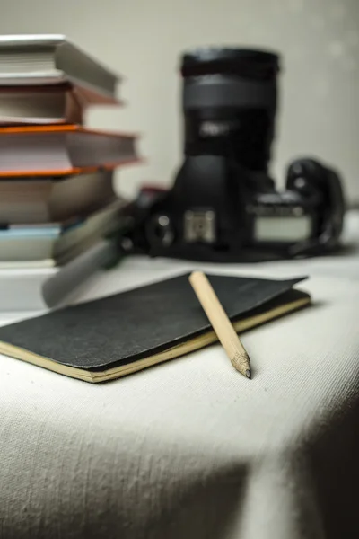 Câmera SLR digital moderna sobre a mesa ao lado de uma pilha de livros, e em primeiro plano um notebook e lápis . — Fotografia de Stock