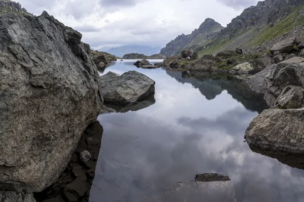 Panoramalandschaft mit einem See in den Bergen, riesigen Felsen und Steinen an der Küste und dem Widerschein von Wolken — Stockfoto