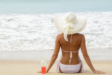 Geniş kenarlı şapkalı kadın elinde soğuk bir kokteyl bardağıyla sahilde oturuyor.