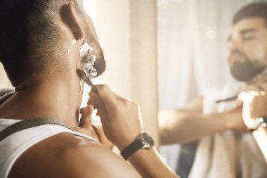 Aynaya bakıp sakalını jiletle tıraş eden yakışıklı bir adam.