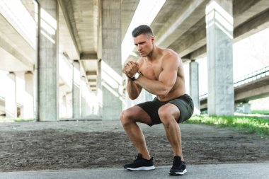 Genç ve kaslı adam, sokak çalışması sırasında köprünün altında çömeliyor.