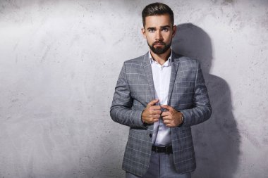 Gri kareli takım giyen yakışıklı sakallı adam beton duvara karşı poz veriyor.