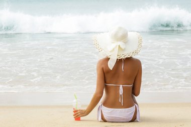 Geniş kenarlı şapkalı kadın elinde soğuk bir kokteyl bardağıyla sahilde oturuyor.
