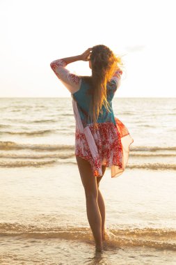 Güzel elbiseli genç bir kadın gün batımında deniz kıyısında yürüyor.