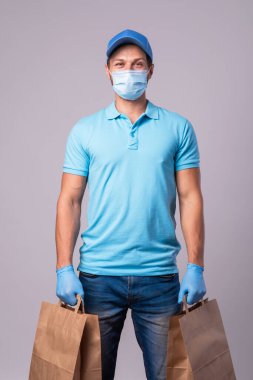 Genç teslimatçı koruyucu maske ve eldivenler takıyor. Gri arka planda izole edilmiş kese kağıtları var.