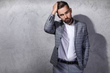 Gri kareli takım giyen yakışıklı sakallı adam beton duvara karşı poz veriyor.