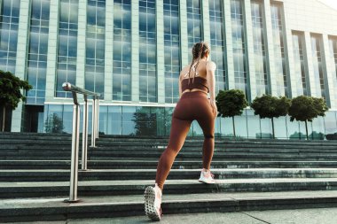 Genç ve sportif bir kadın üst katta beton bir merdivenin yanında koşuyor.