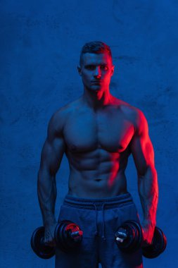 Genç ve kaslı vücut geliştirici adam dambıllarla çalışıyor.