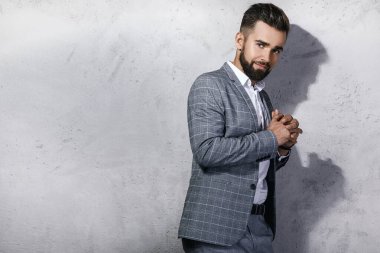 Gri kareli takım giyen yakışıklı sakallı adam beton duvara karşı poz veriyor.