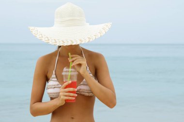 Geniş kenarlı şapkalı kadın sahilde ferahlatıcı bir kırmızı kokteyl içiyor.