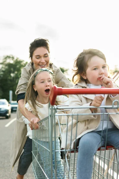 若い幸せな母親と彼女のかわいい娘たちは スーパーマーケットの横にある駐車場でショッピングカートで楽しんでいます — ストック写真