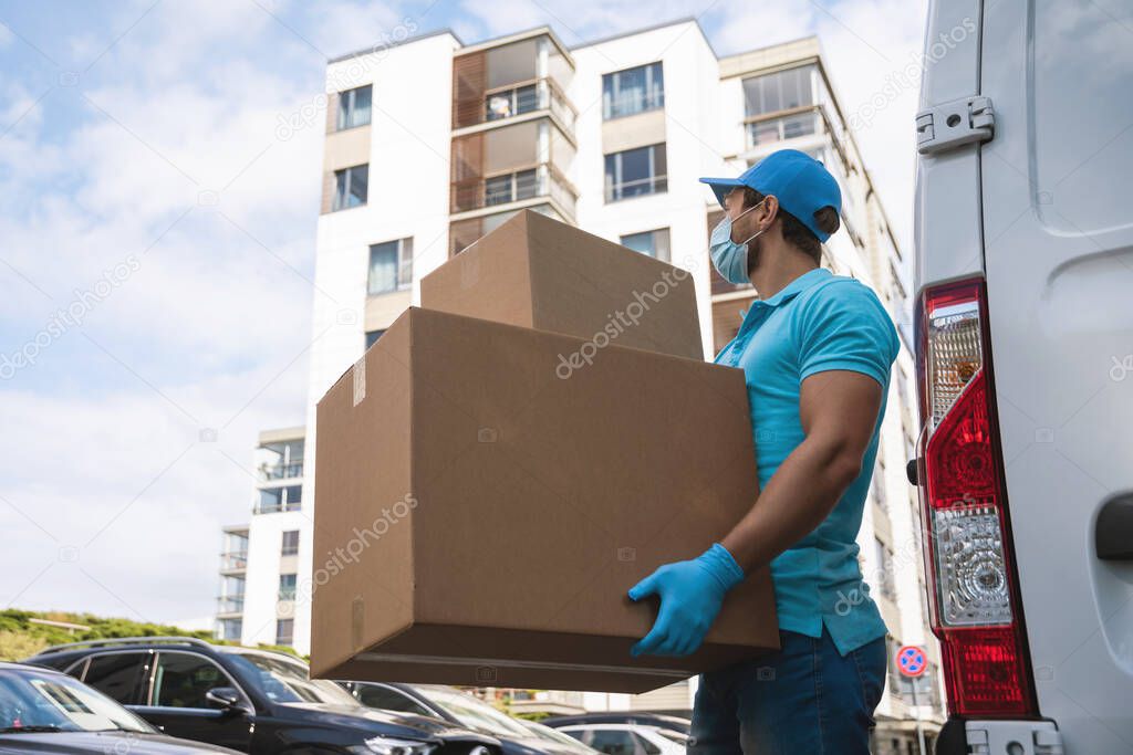 Delivery man wearing prevention mask and gloves is holding stack of the cardboard boxes near his van