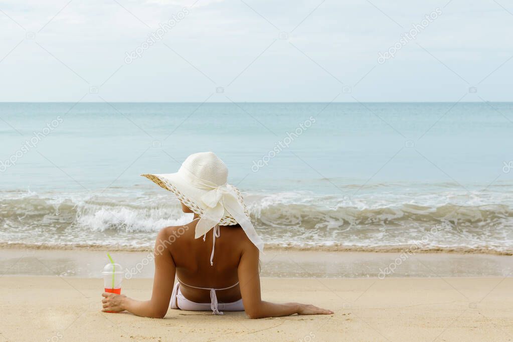 Woman wearing broad-brimmed hat is holding a glass of cold cocktail and sitting on the beach