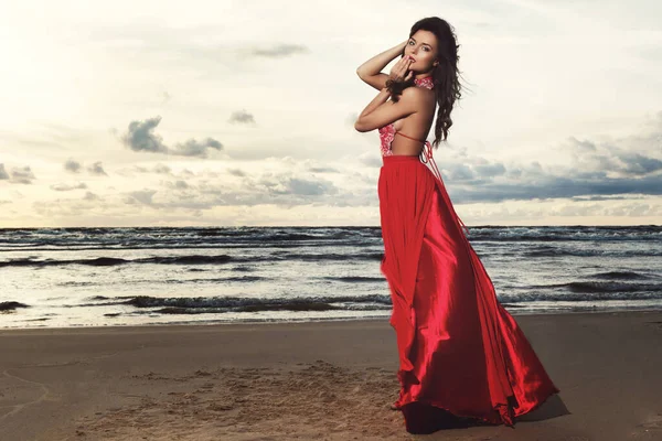 Stunning woman wearing beautiful red dress on the beach during sunset time