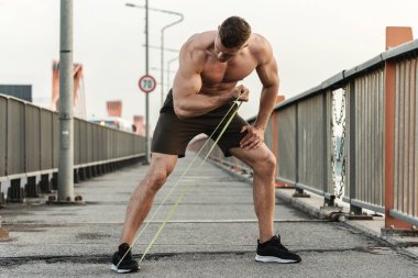 Genç ve kaslı bir adam sokakta dirençli lastik bantlarla çalışırken. Biceps kıvırcık egzersizi.