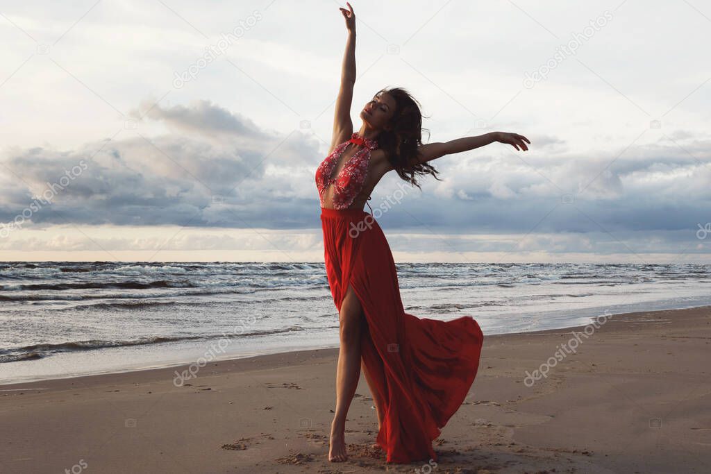 Stunning woman wearing beautiful red dress on the beach during sunset time