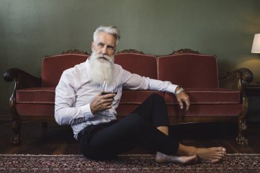 Şık ve yakışıklı sakallı son sınıf öğrencisi yerde oturmuş kırmızı şarap içiyor.