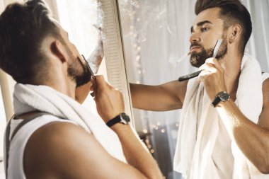 Yakışıklı bir adam aynaya bakıyor, sigara içiyor ve sakalını jiletle tıraş ediyor.