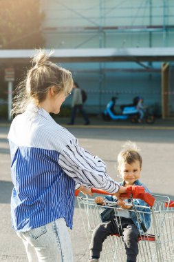 Genç, güzel anne ve küçük tatlı oğlu süpermarketin yanındaki alışveriş arabasıyla eğleniyorlar.
