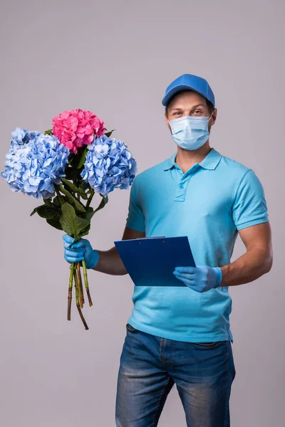 Young delivery man with a bouquet of flowers is wearing prevention mask and gloves isolated on gray background