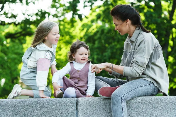 若い幸せな母親と彼女の2人の娘が座って街の公園で遊んでいます — ストック写真