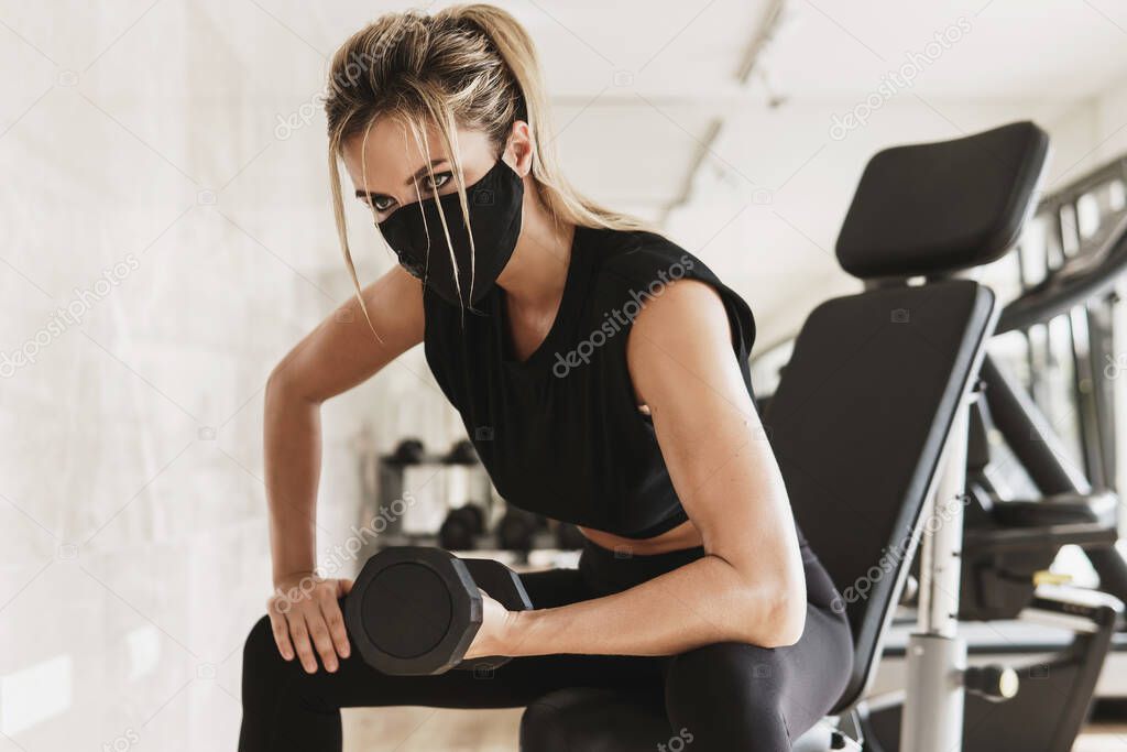 Gym new normal. Young athletic woman wearing a prevention face mask during her fitness workout with a dumbbells.