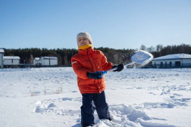 Kışın giyinmiş bir çocuk soğuk havada güneşli bir günde banliyödeki evlerin yakınlarında kar kürüyor..
