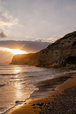 Engebeli bir kıyı manzarasının üzerinde gün batımı deniz dalgaları ve kayalık kıyı şeridine altın ışık saçıyor.. 
