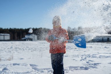 Kışın giyinmiş bir çocuk soğuk havada güneşli bir günde banliyödeki evlerin yakınlarında kar kürüyor..