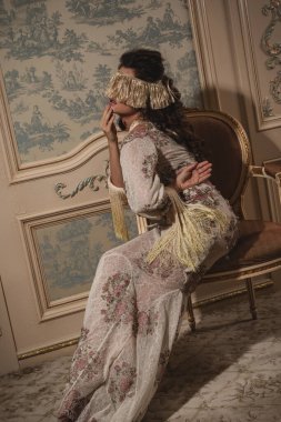 Karmaşık dantel bir elbise giymiş bir kadın lüks bir iç mekanda zarafetle poz veriyor..