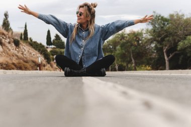 Kot gömlek giyen neşeli genç bir kadın boş bir kırsal yolda oturuyor..