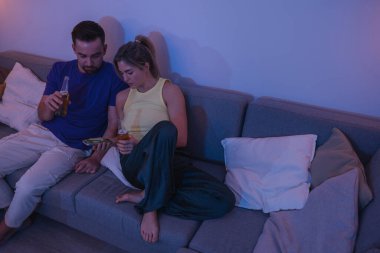  Evdeki rahat bir akşamda içki ve akıllı telefonla koltukta oturan çift..