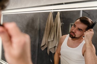 Modern bir banyoda aynaya bakarken saçını tarayan bir adam..