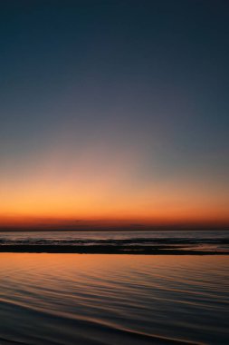 Gün batımında sakin deniz manzarası. Canlı turuncu ve mavi eğimli gökyüzü sakin okyanus dalgalarını yansıtıyor. Huzurlu sahil manzarası.