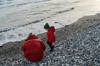 Annesi ve oğlu, kalın ceketler giyip, çakıllı bir plajda farklı taşlar toplayıp incelerler..