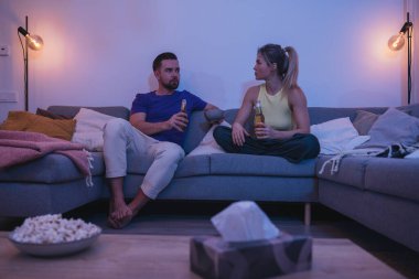 Çift kanepede oturuyor, sıcak bir oturma odasında bira ve nachos yiyor..