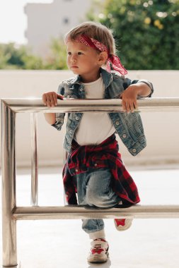 Kot elbise giyen ve açık hava ortamında metal korkuluklarda kırmızı bandana çalan tatlı çocuk..