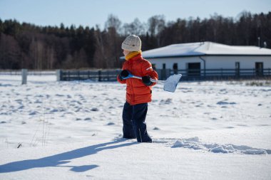 Kışın giyinmiş bir çocuk soğuk havada güneşli bir günde banliyödeki evlerin yakınlarında kar kürüyor..