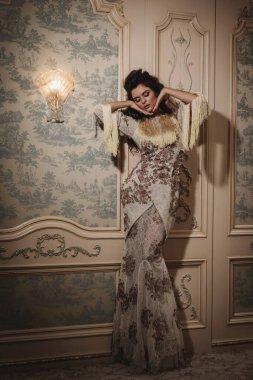 Karmaşık dantel bir elbise giymiş bir kadın lüks bir iç mekanda zarafetle poz veriyor..