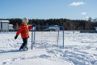 Karla kaplı bahçede kale direğiyle futbol oynayan, kışlık kıyafetler giyen, dışarıda eğlenen ve mevsimlik aktivitelerin tadını çıkaran bir çocuk..