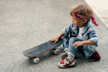 Kot elbise giyen, kırmızı bandana takan küçük çocuk şehir ortamında kaykayla oynuyor..