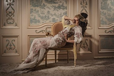 Karmaşık dantel bir elbise giymiş bir kadın lüks bir iç mekanda zarafetle poz veriyor..