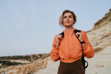 Turuncu kapüşonlu ve siyah sırt çantalı genç bir kadın engebeli kıyı manzarasını keşfediyor..