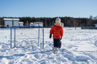 Karla kaplı bahçede kale direğiyle futbol oynayan, kışlık kıyafetler giyen, dışarıda eğlenen ve mevsimlik aktivitelerin tadını çıkaran bir çocuk..