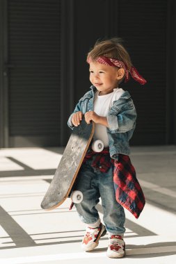 Kot elbise giyen, kırmızı bandana takan küçük çocuk şehir ortamında kaykayla oynuyor..