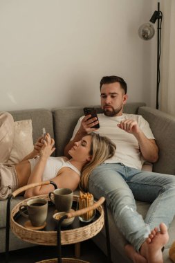 Çift, yakınlarda atıştırmalık eşliğinde akıllı telefonlar kullanarak koltukta dinleniyor..