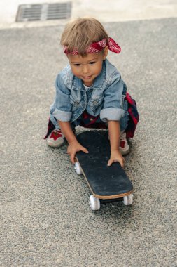 Kot elbise giyen, kırmızı bandana takan küçük çocuk şehir ortamında kaykayla oynuyor..