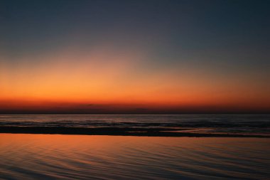 Gün batımında sakin deniz manzarası. Canlı turuncu ve mavi eğimli gökyüzü sakin okyanus dalgalarını yansıtıyor. Huzurlu sahil manzarası.